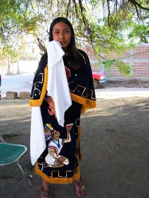Young woman wearing a traditional embroidered dress with a white cloth over her shoulder, standing outdoors under a tree.