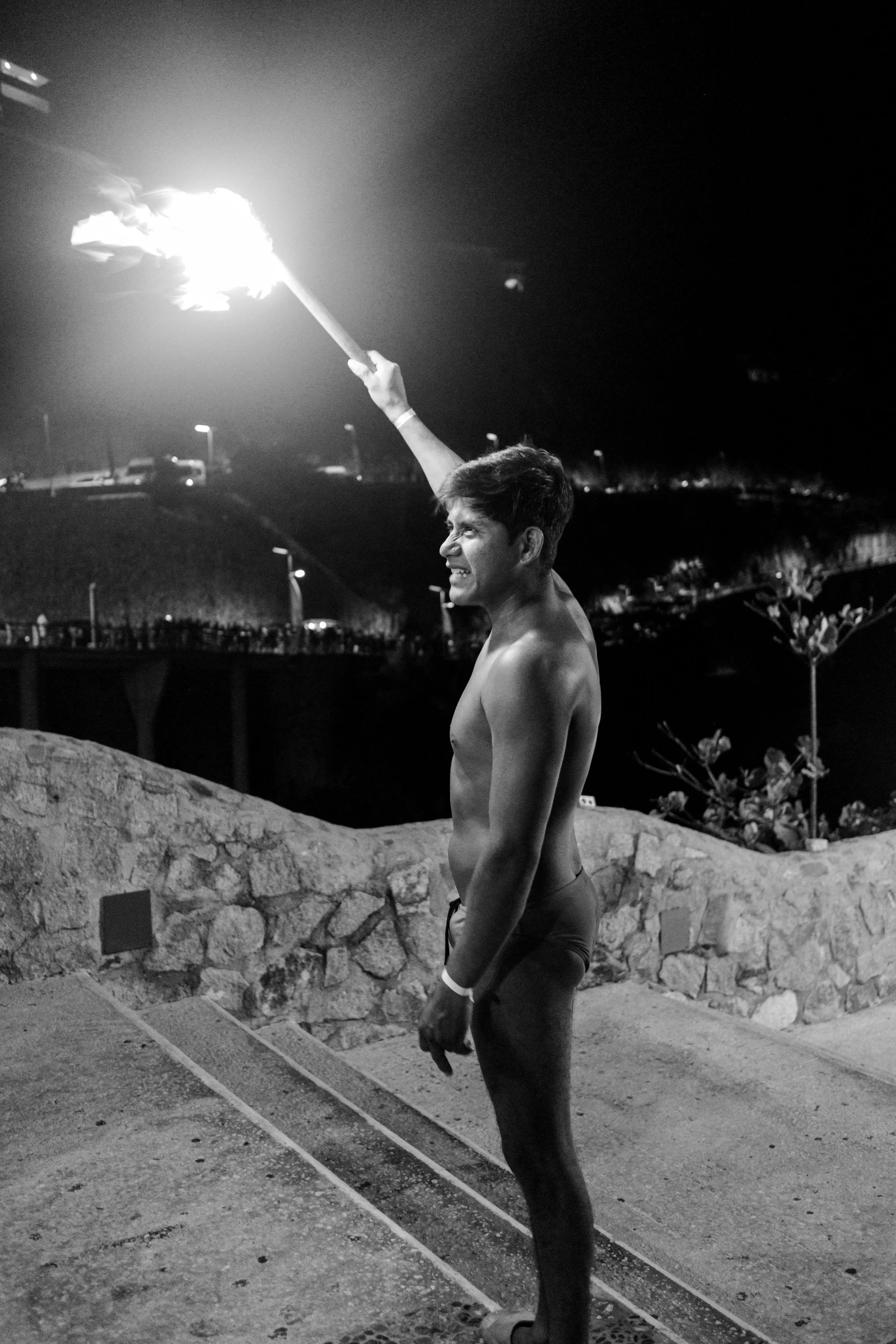 A young man standing shirtless outdoors at night holding a burning torch with flames. He appears to be smiling and relaxed, with a stone wall and illuminated streetlights in the background.