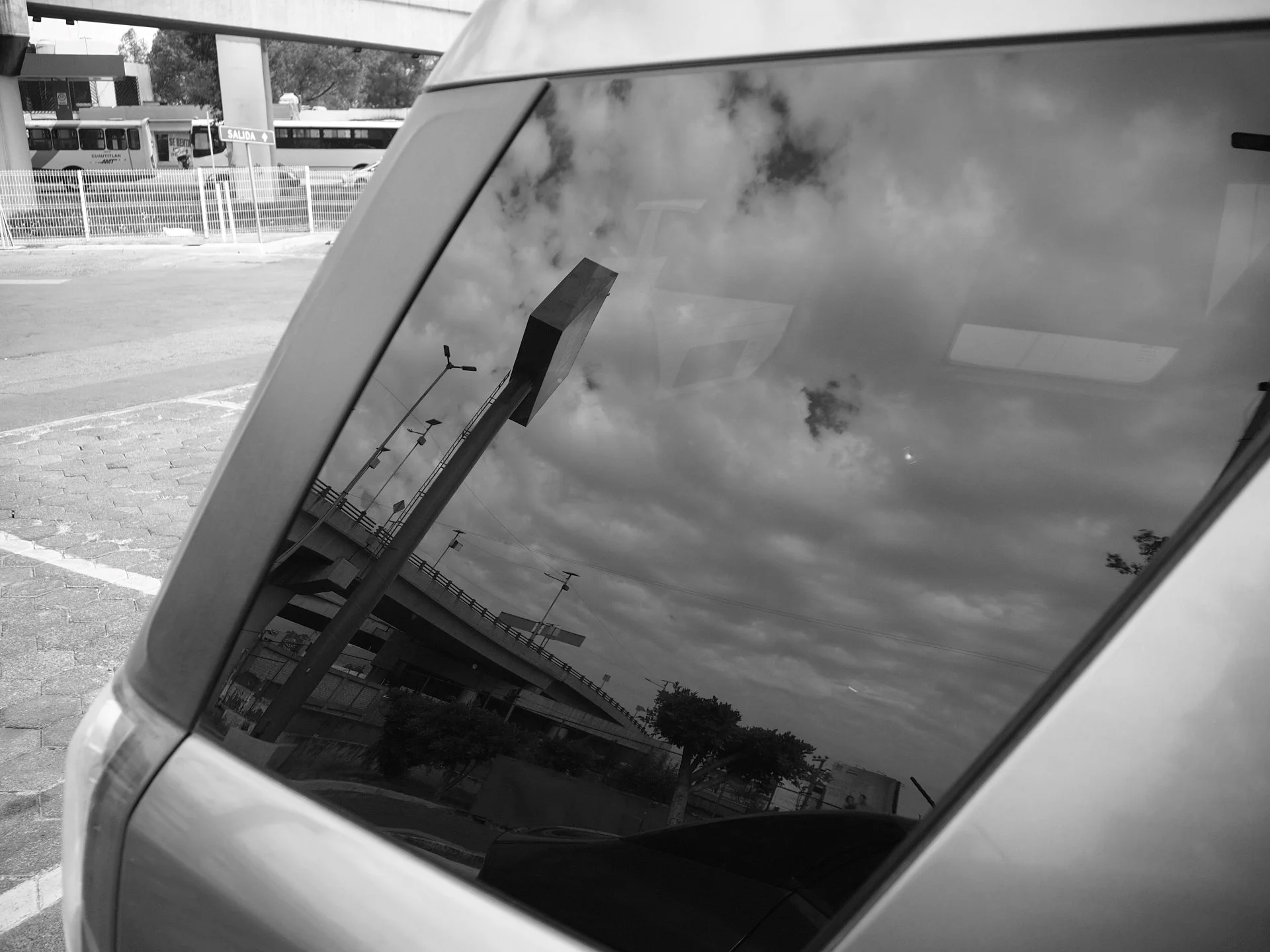 Reflection of a bridge, lamp posts, trees, and a cloudy sky in the side window of a vehicle.