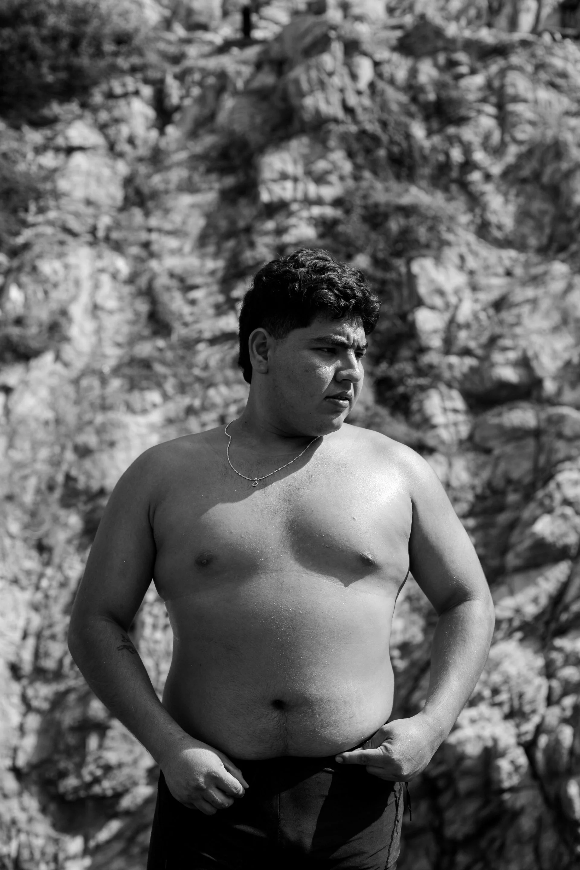 A shirtless young man with short dark curly hair standing outdoors in front of a rocky background, with his left arm bent and hand on his hip, wearing a necklace.