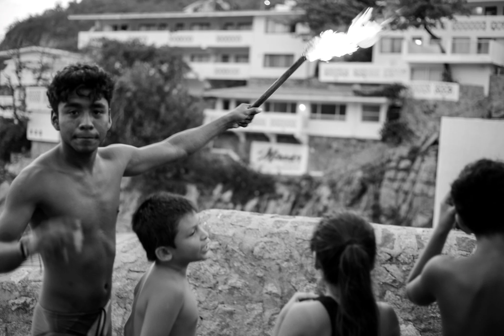 A shirtless young man holding a lit stick with flames extending from it, surrounded by children outdoors, with buildings and trees in the background.