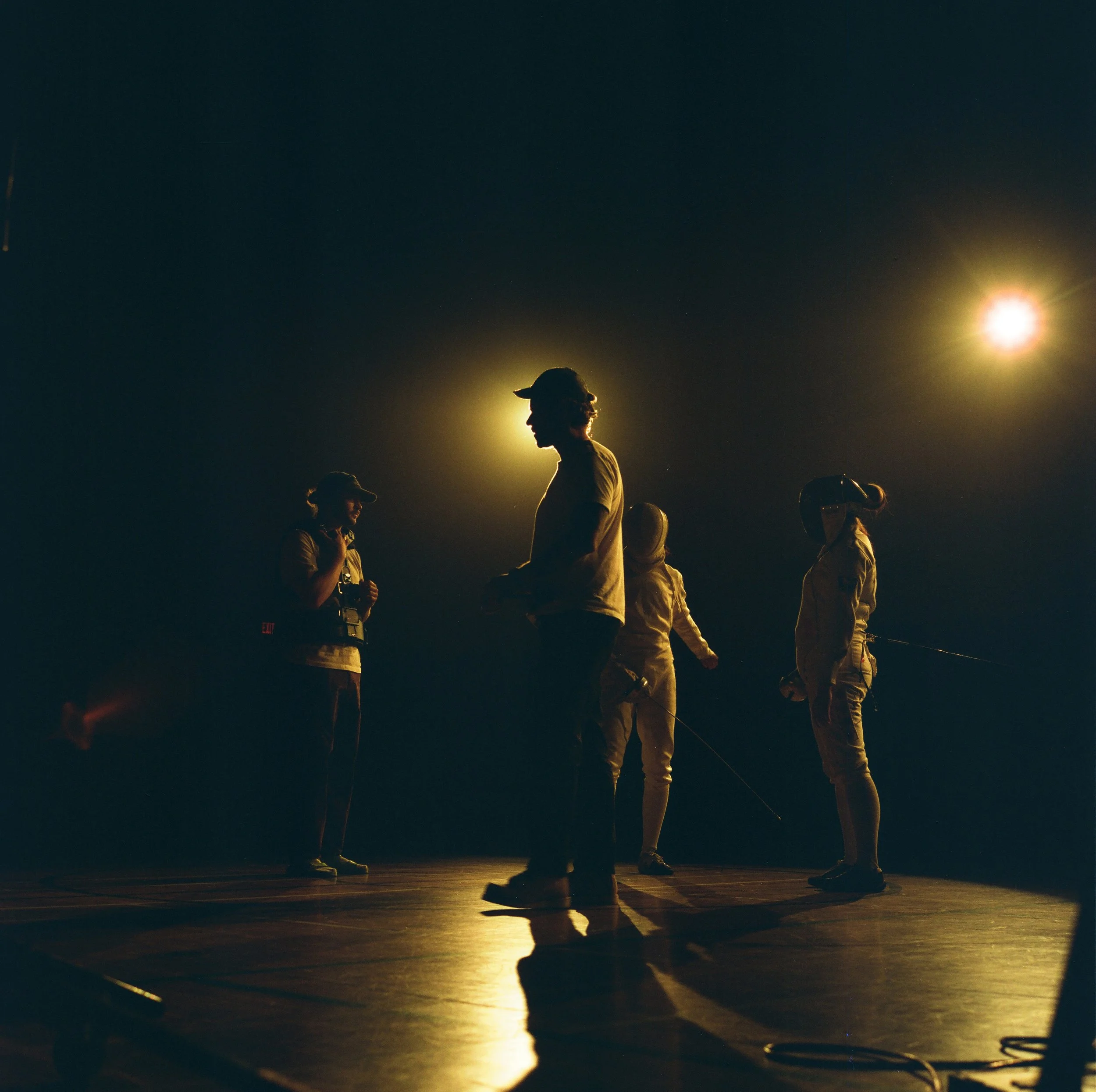 Silhouettes of four people on stage under yellow lights, some wearing hats and protective gear, with a dark background.