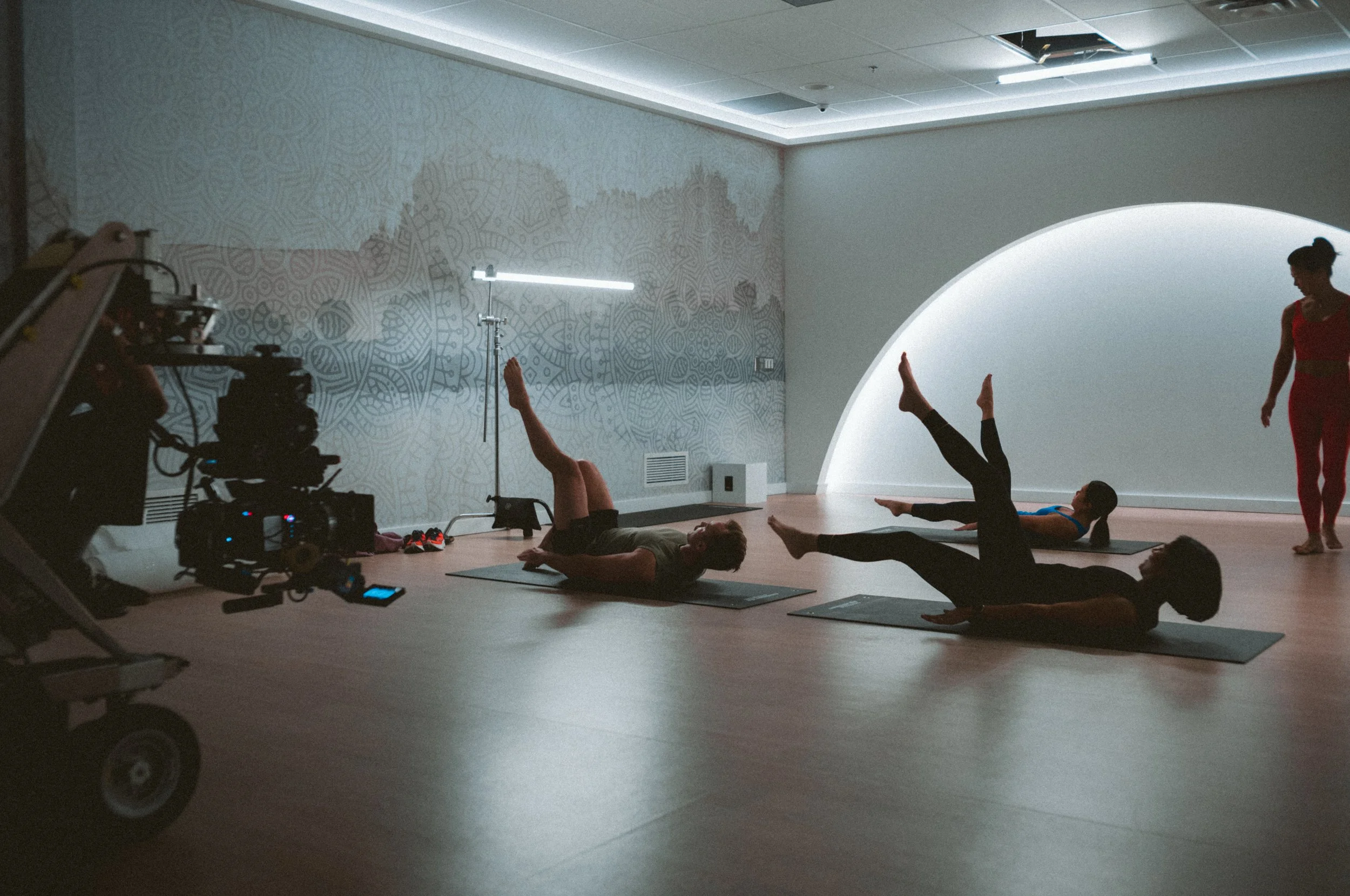 Group of people practicing yoga in a dimly lit room, with a camera set up nearby for recording or filming.