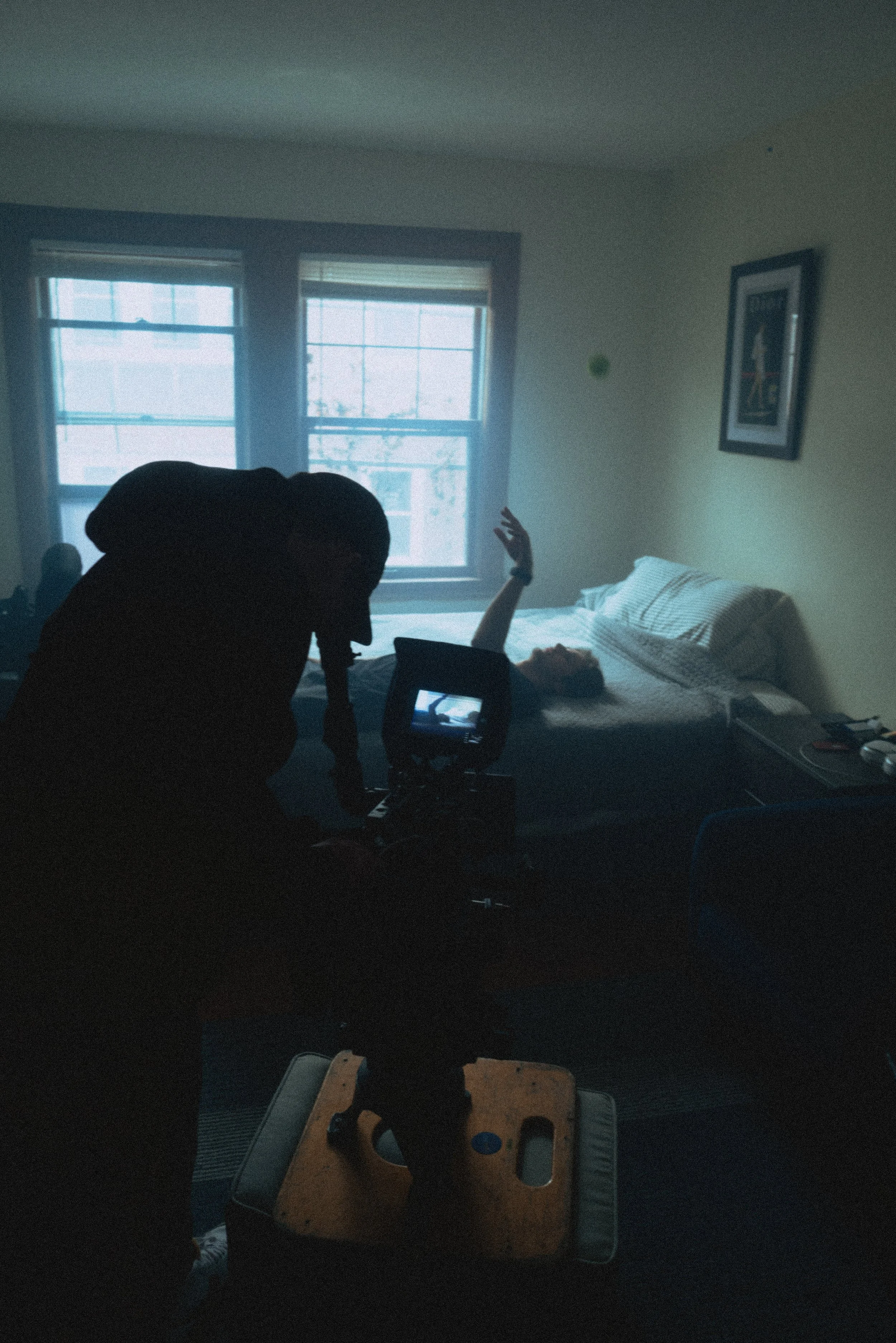 A person lying in bed with their arm raised as a videographer films from a low angle in a dimly lit room with two windows and artwork on the wall.