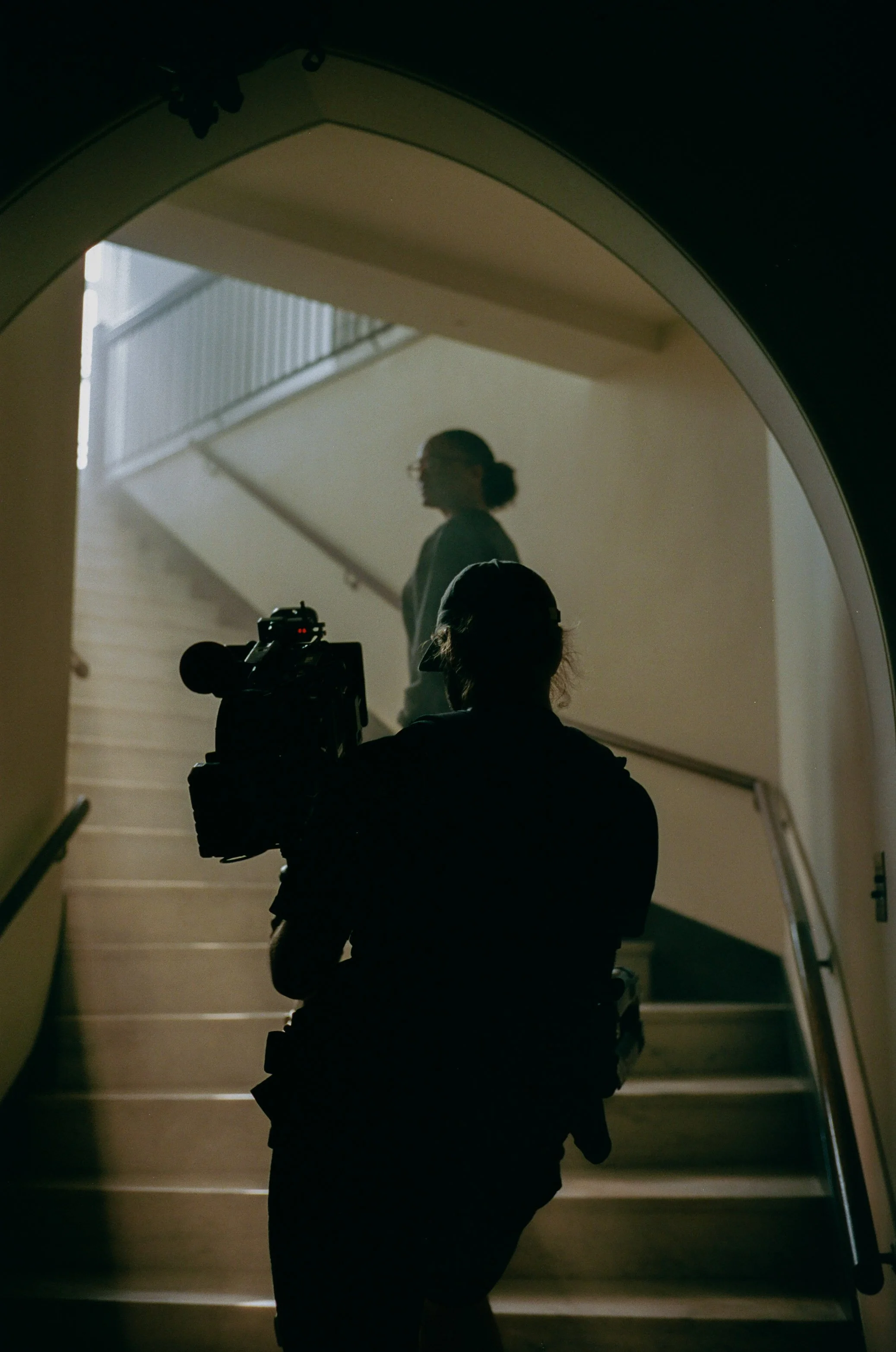 Silhouette of a person holding a camera, standing at the bottom of a staircase, with a woman walking up the stairs in the background.