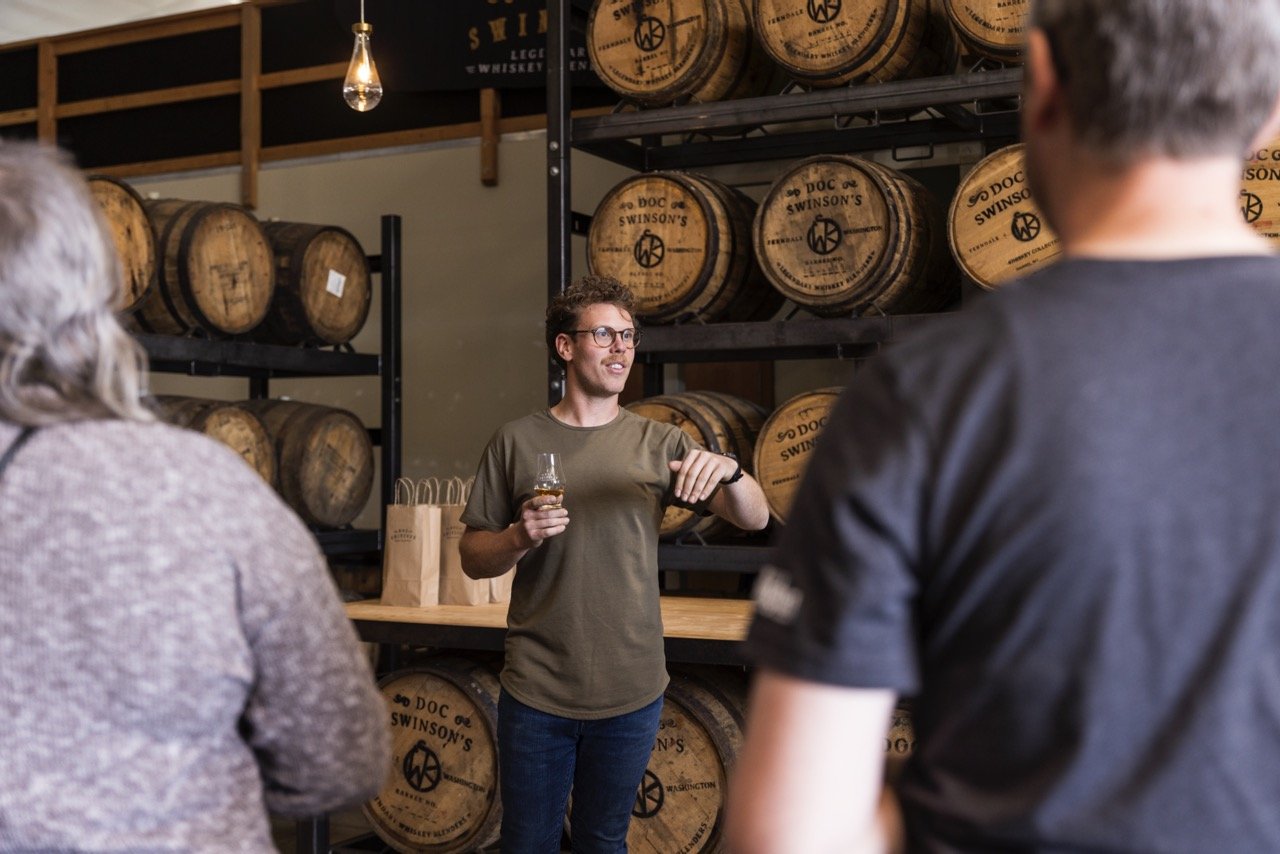 A man giving a whiskey tasting or tour to a small group in a whiskey warehouse, with barrels on shelves in the background.