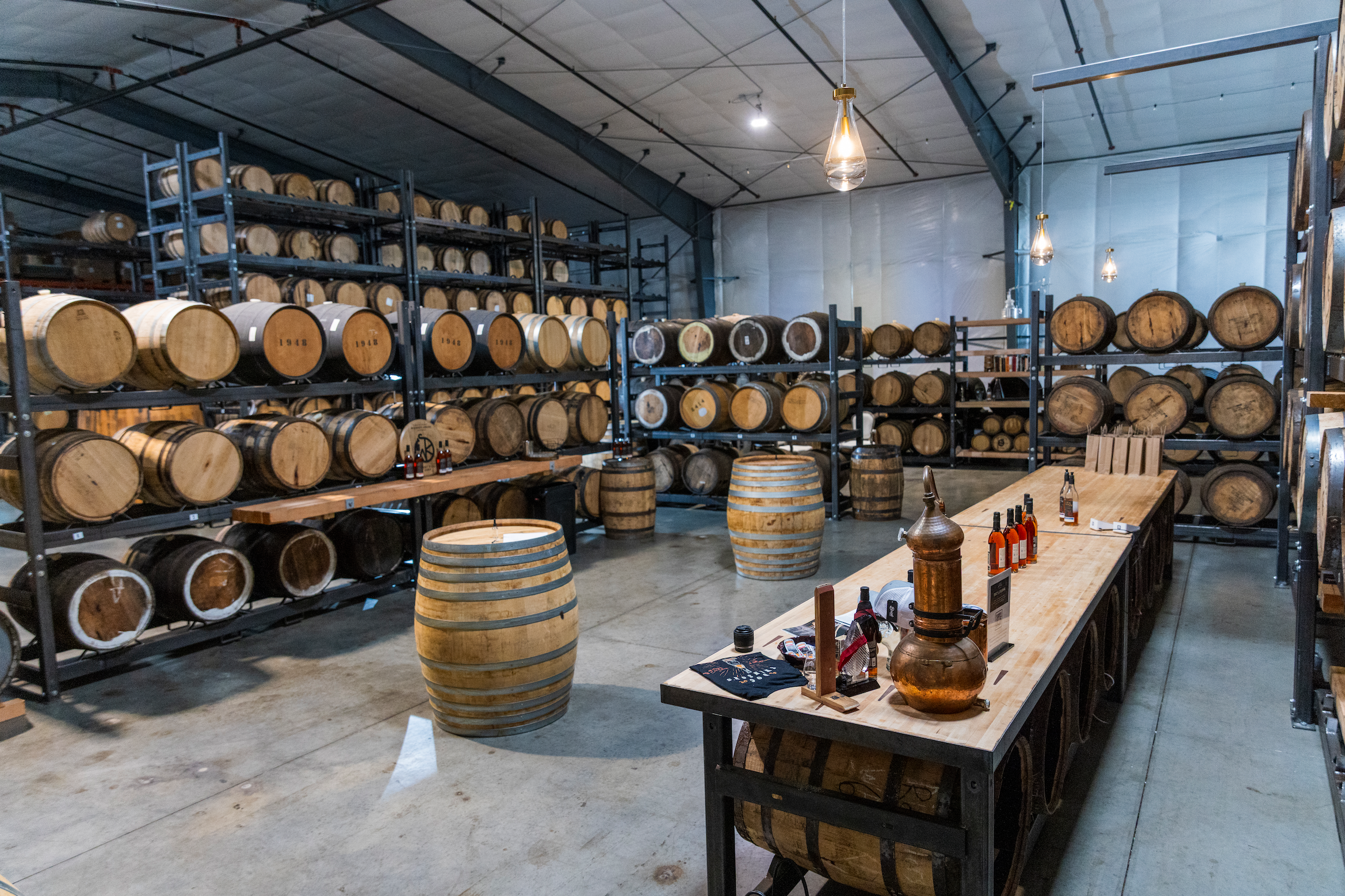 A warehouse with shelves filled with wooden barrels, possibly for aging spirits like whiskey or bourbon, and a table with tasting bottles and equipment, suggesting a whiskey tasting or distillery setting.