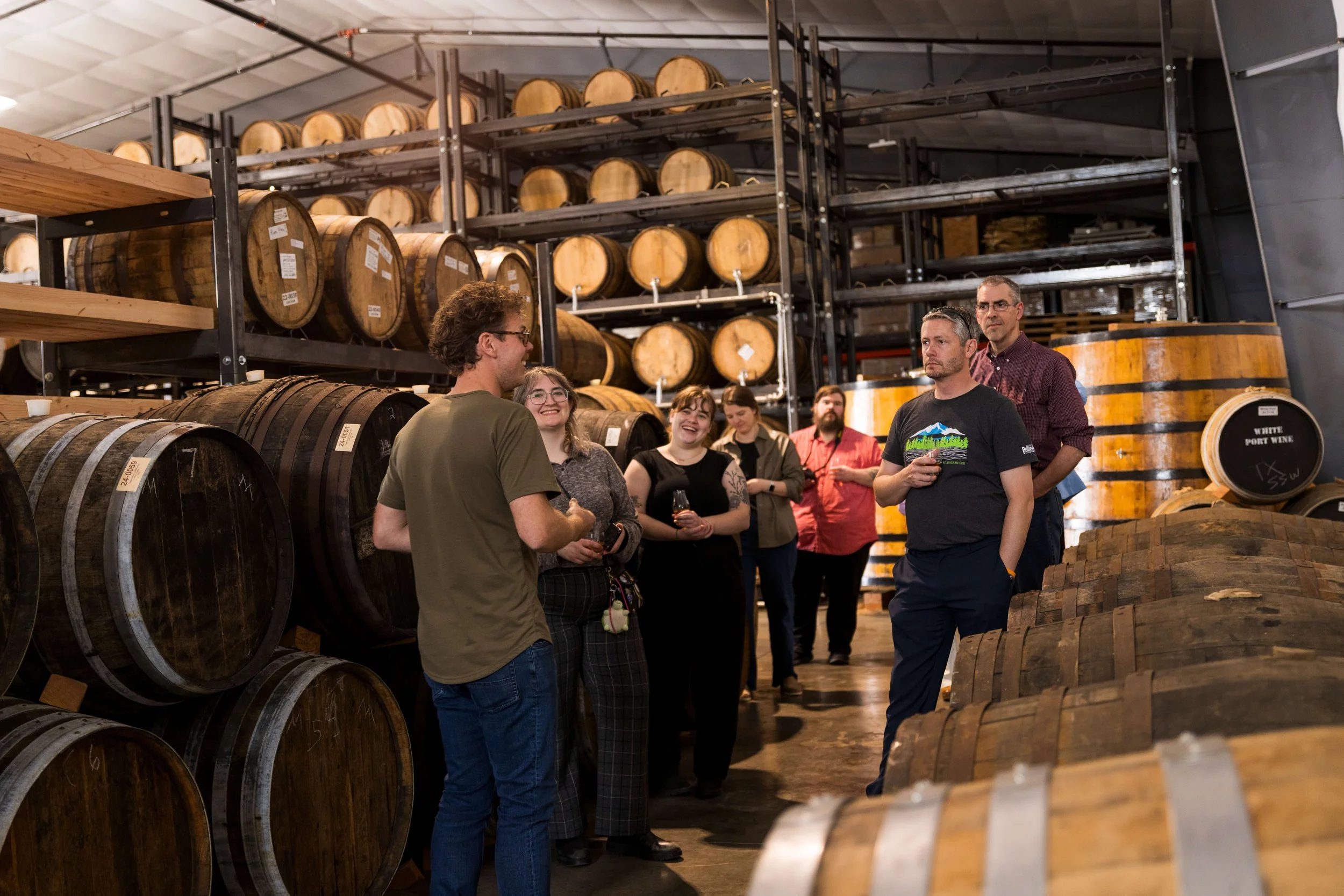 A group of people in a wine barrel storage warehouse, some holding wine glasses, engaging in conversation with a guide.