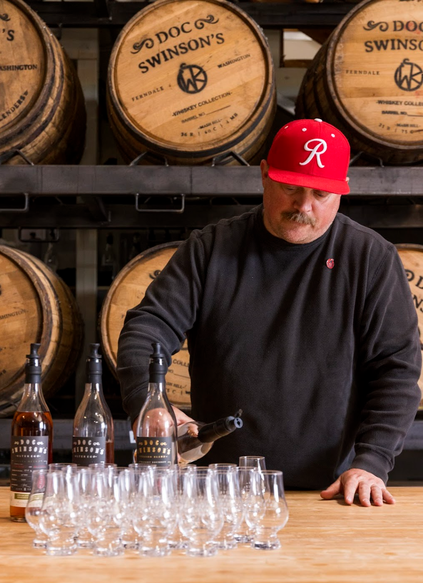 A man in a black shirt and red baseball cap preparing whiskey at a distillery. There are whiskey bottles and glasses on the wooden table, and large whiskey barrels in the background.