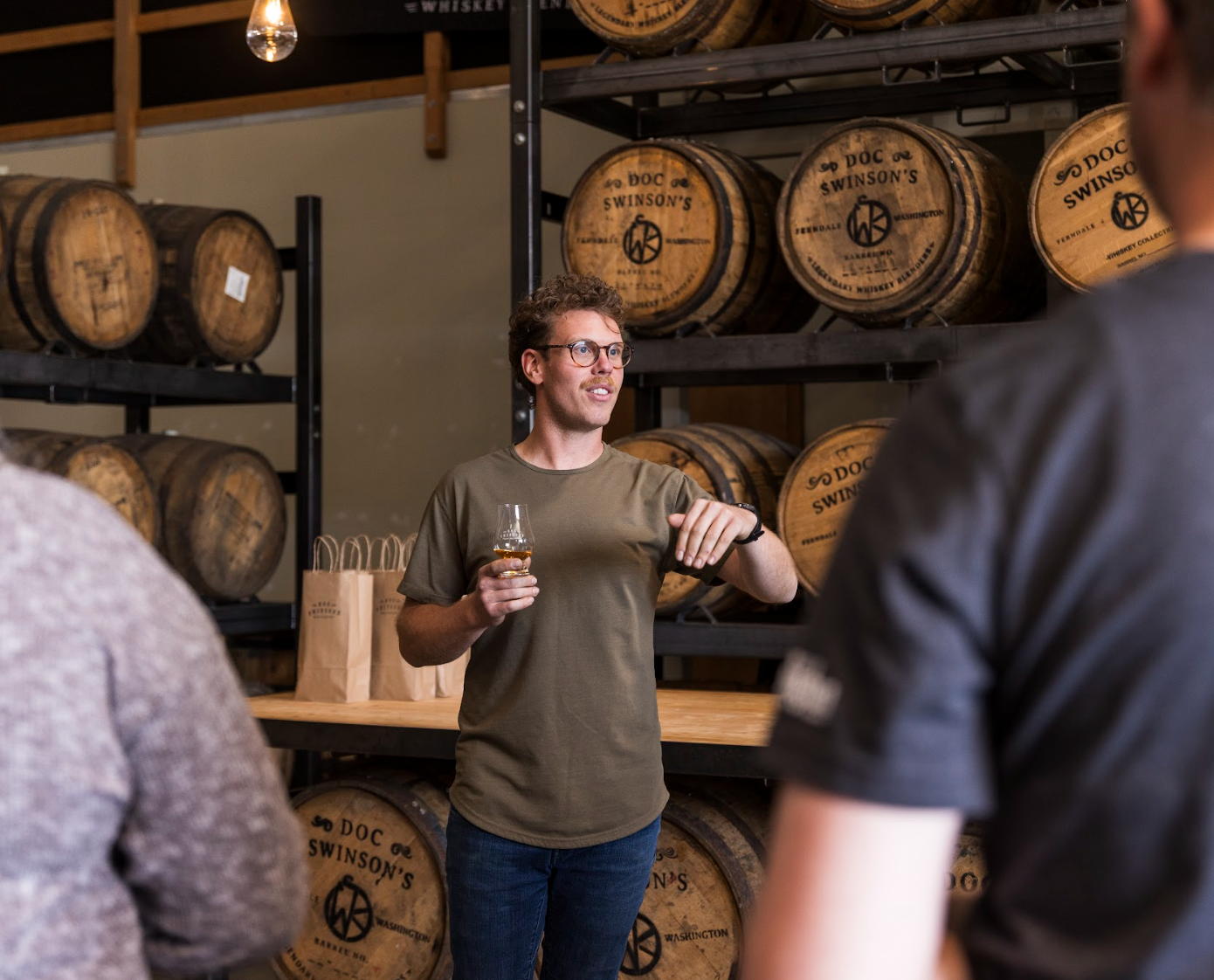 A man giving a presentation in a whiskey warehouse with barrels on shelves behind him, holding a glass of whiskey, while three people listen.