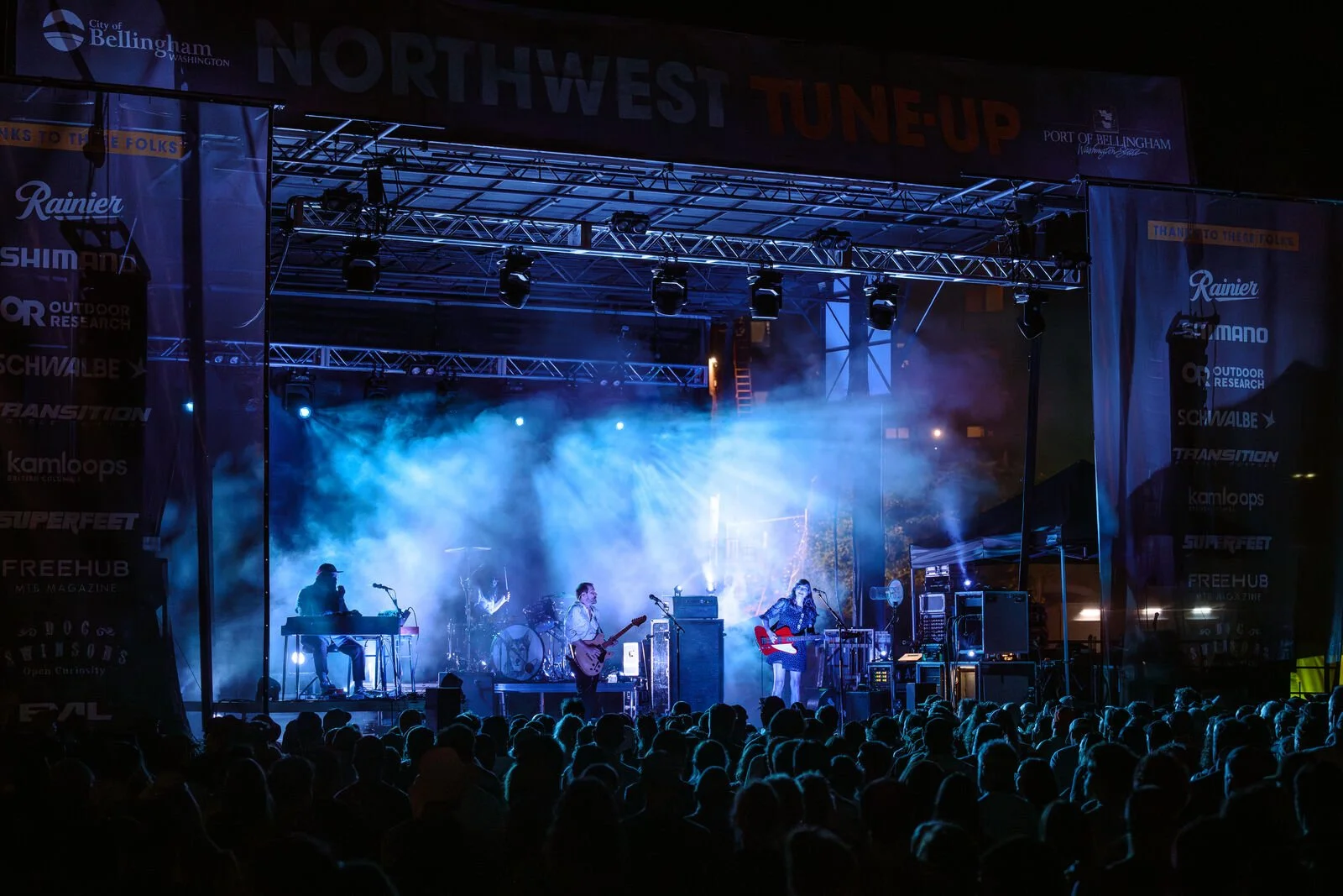 A band performing on an outdoor stage at night, with blue and white stage lights and smoke effects, and a large audience in front of the stage.