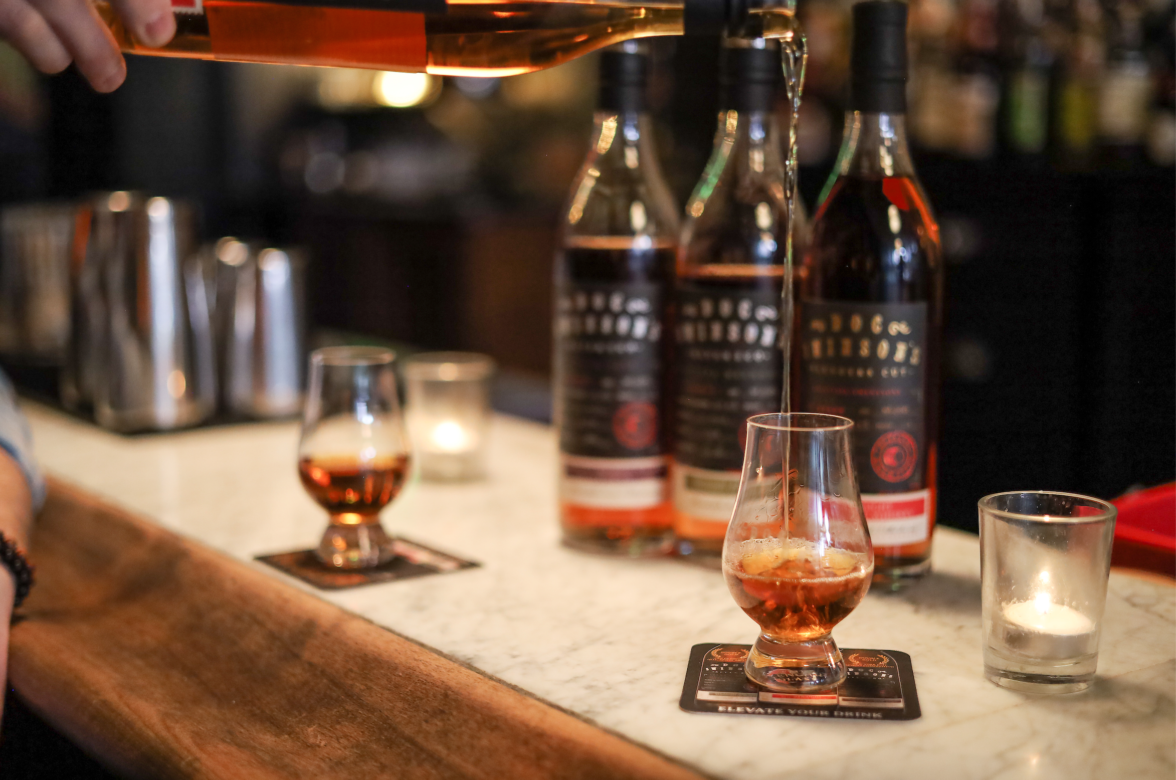 A bartender pours a dark beverage into a short glass on a bar counter. There are three bottles of alcohol behind the glass, and small candles are also on the counter, creating a cozy atmosphere.