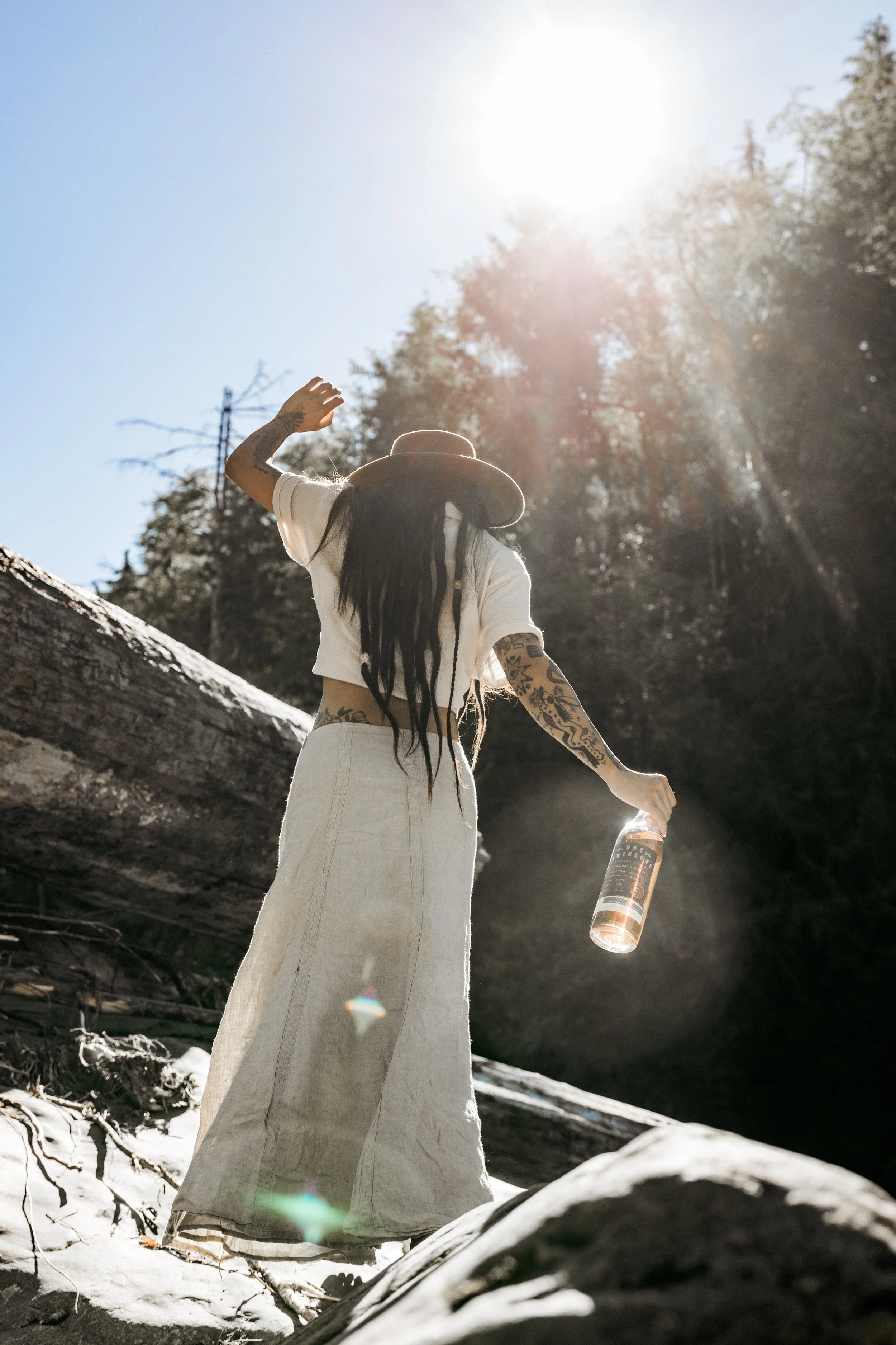 A woman with tattoos in wide-leg pants and a cropped shirt, wearing a hat, stands outdoors on a sunny day, holding a bottle of rosé wine, with a fallen log and trees in the background.
