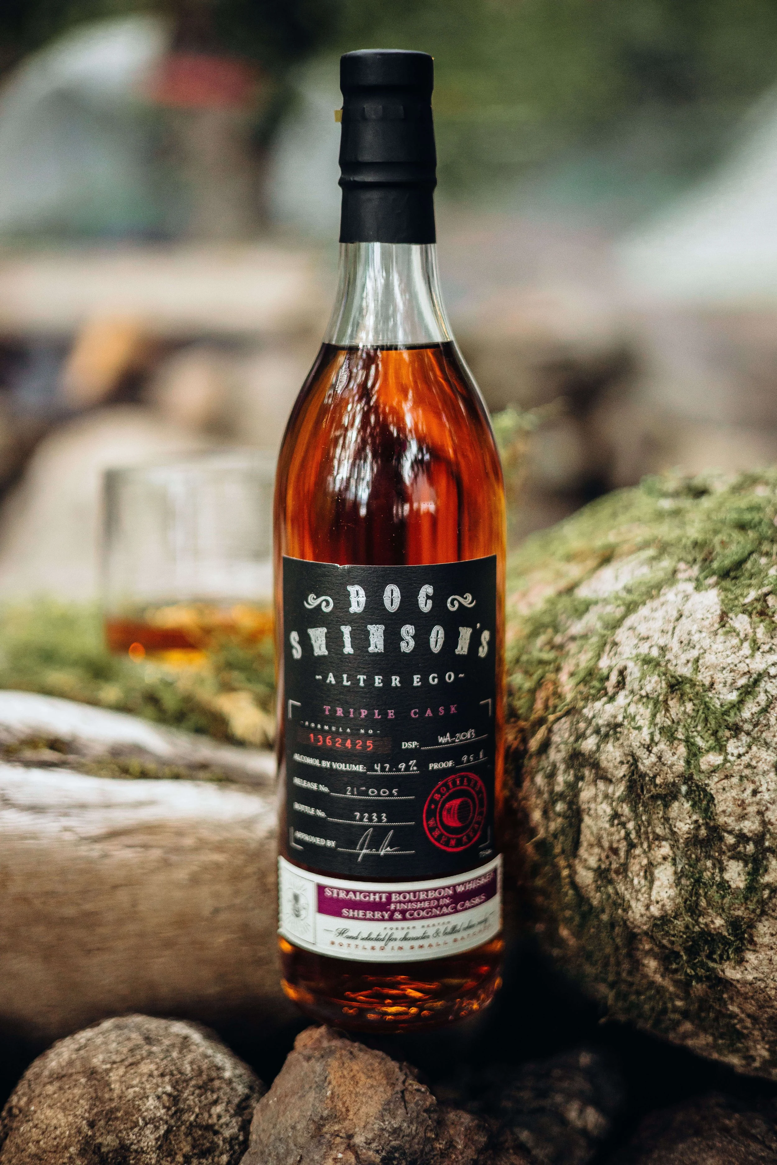 Bottle of Dog Stillson's Alter Ego Triple Cask whiskey on rocks surrounded by mossy rocks and a blurred background.