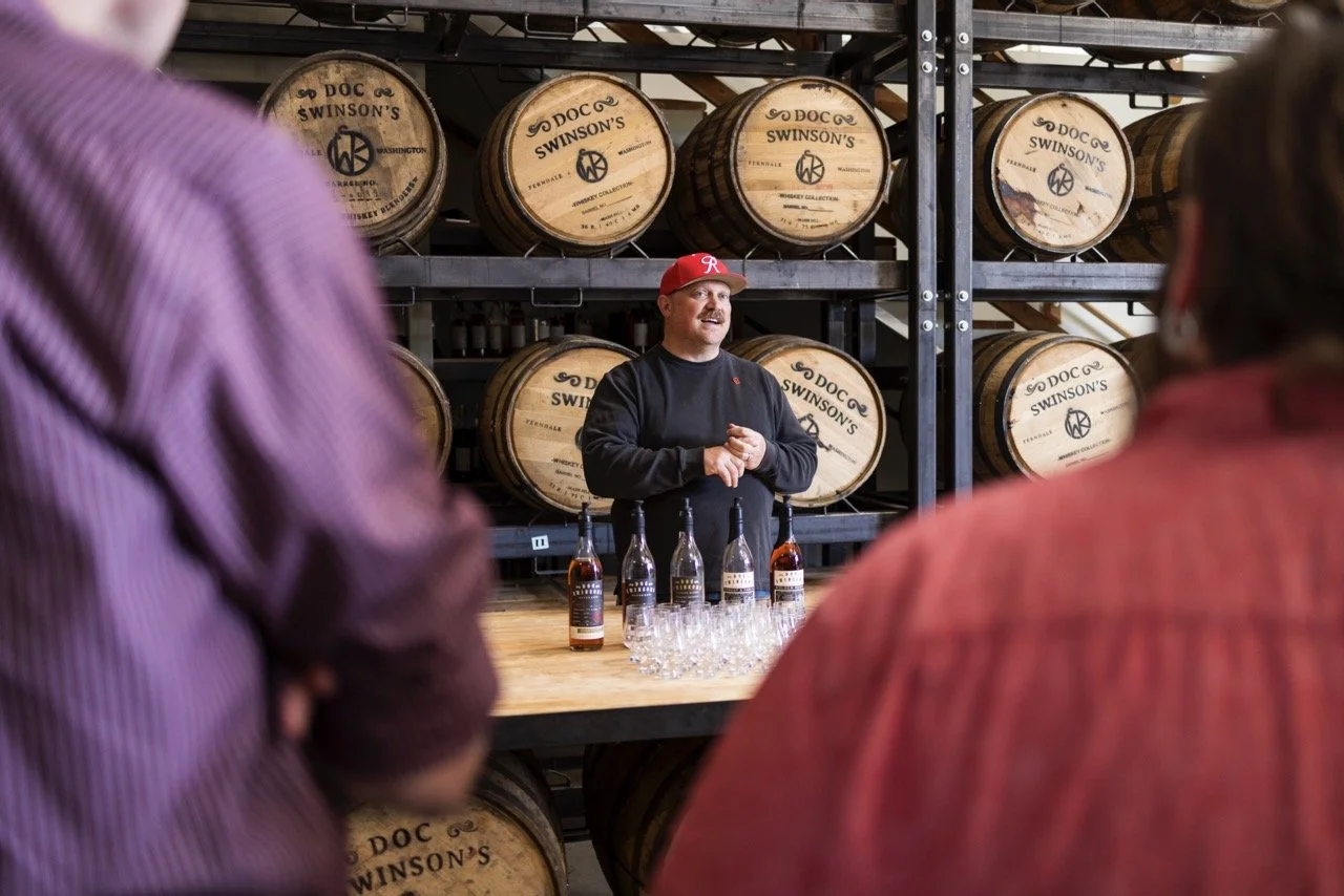 A man with a red cap and black sweatshirt giving a whiskey tasting presentation in front of whiskey barrels labeled 'Doc Swinson's.'" There are bottles and glasses on the table in front of him, and two audience members are partially in view.
