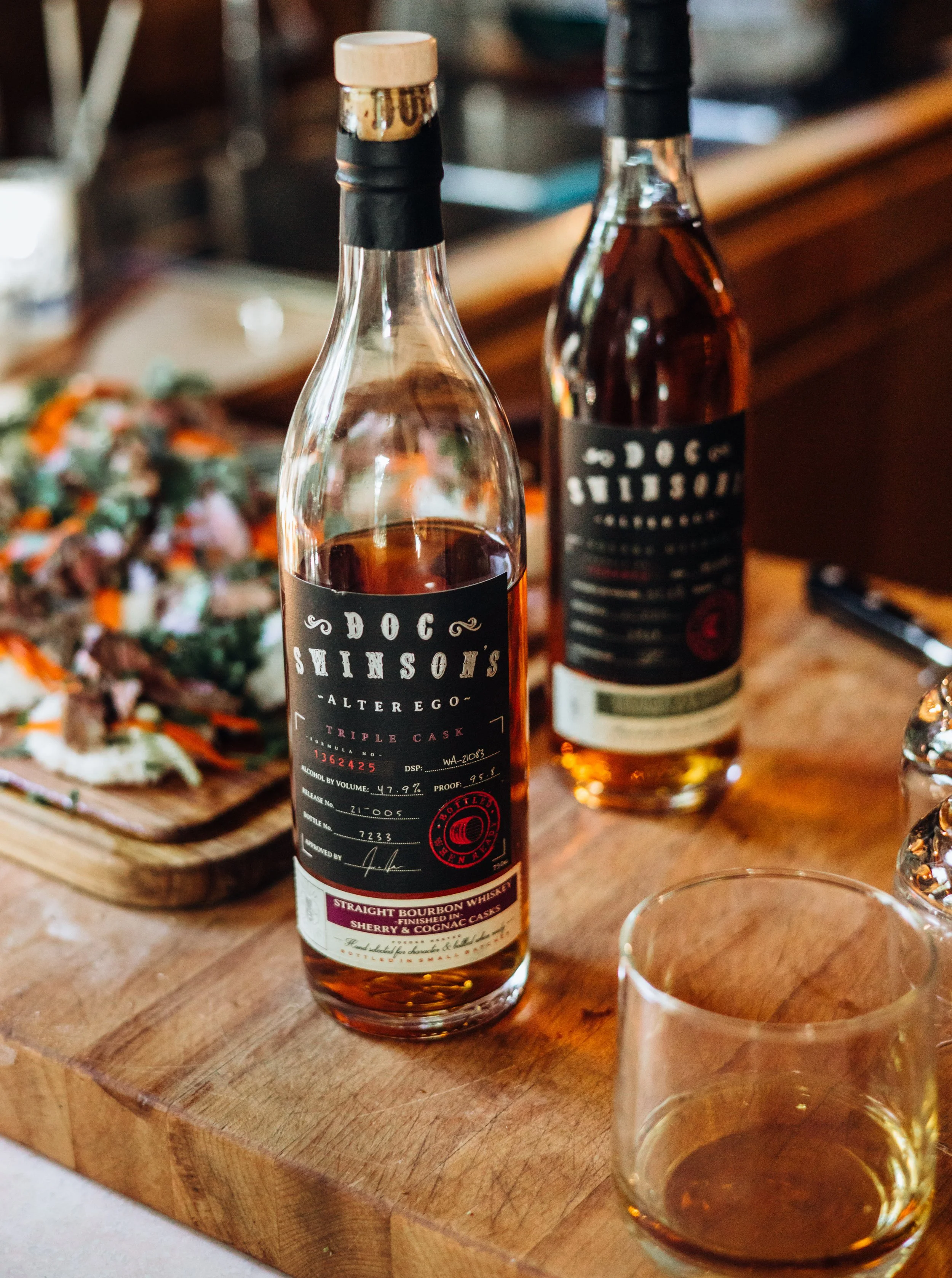 Two bottles of DOC Sunson's alter ego whiskey on a wooden table, one open with a glass of whiskey nearby, and a salad on a wooden plate in the background.
