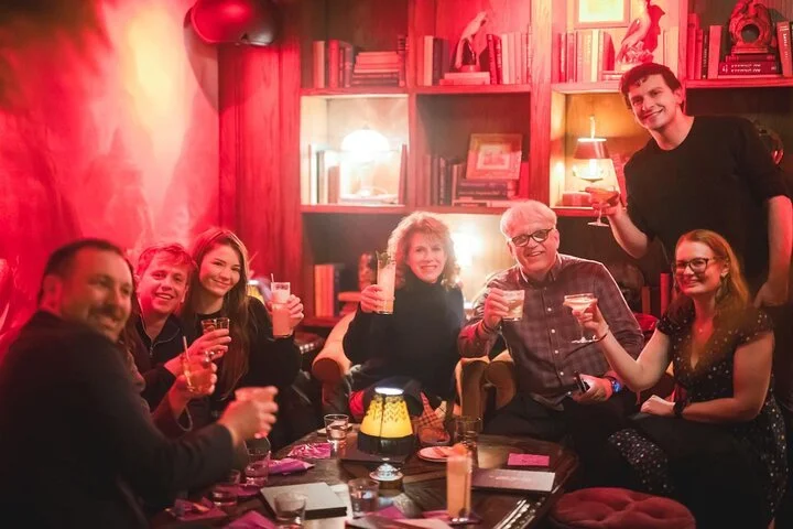 Group of eight people sitting and standing around a table in a cozy, warmly lit room, enjoying drinks and smiling at the camera.