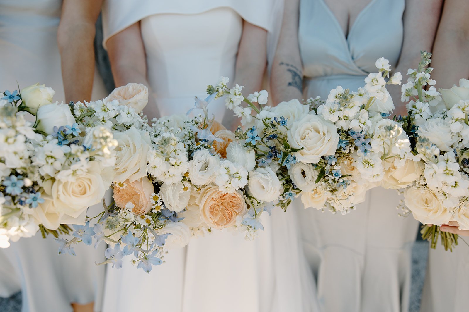 Wedding bouquet made of white and light peach roses, blue delphiniums, and white baby's breath held by bridal party members in white dresses.