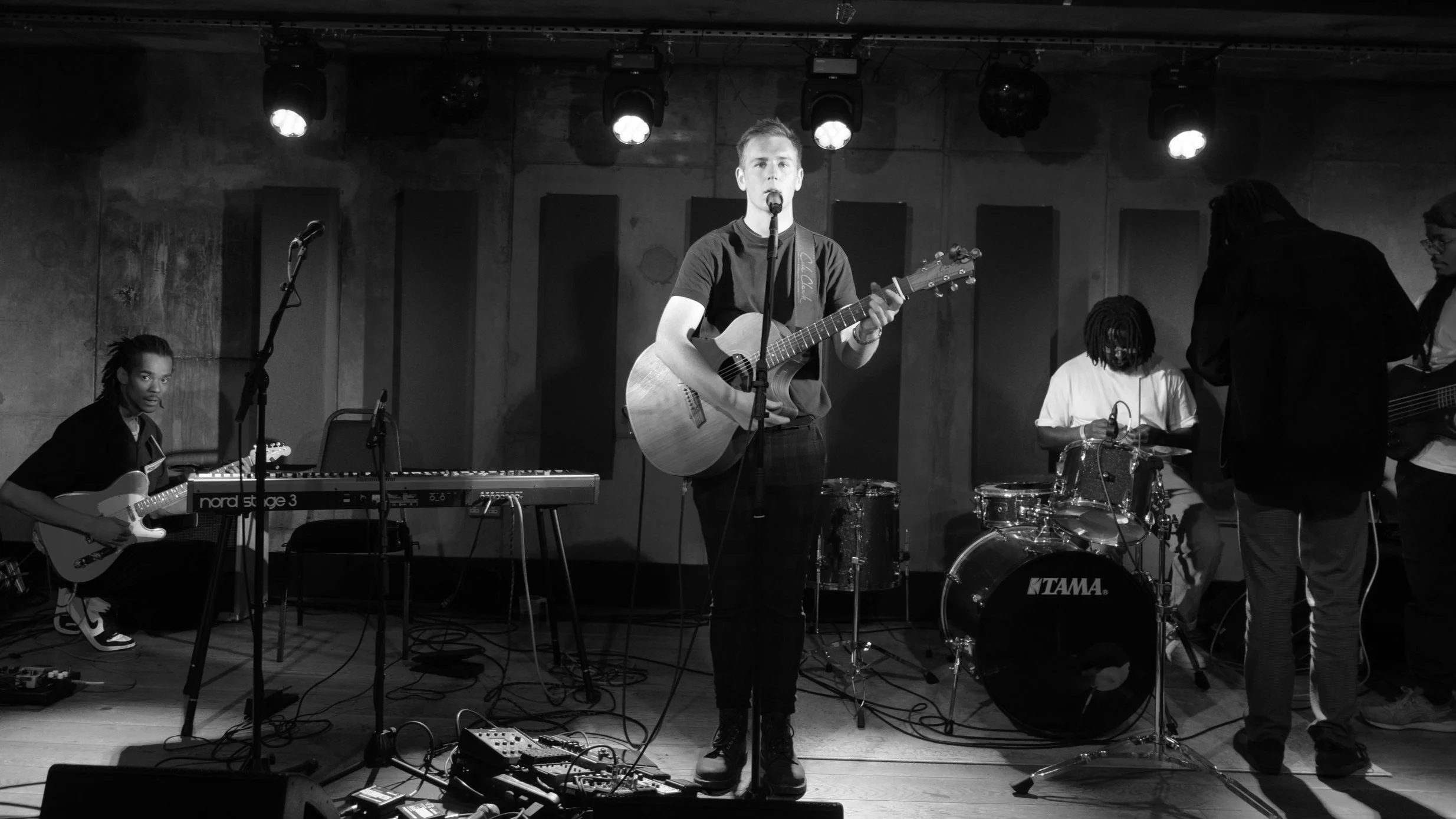 Black and white photo of a band performing on stage, featuring a guitarist on acoustic guitar, a keyboard player, a drummer at a drum kit, and a bassist.