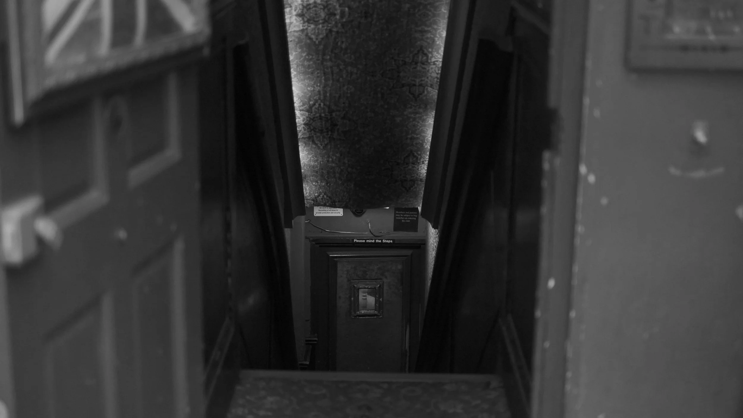 Narrow staircase in dimly lit hallway with patterned carpet and warning sign reading "Please mind the steps."