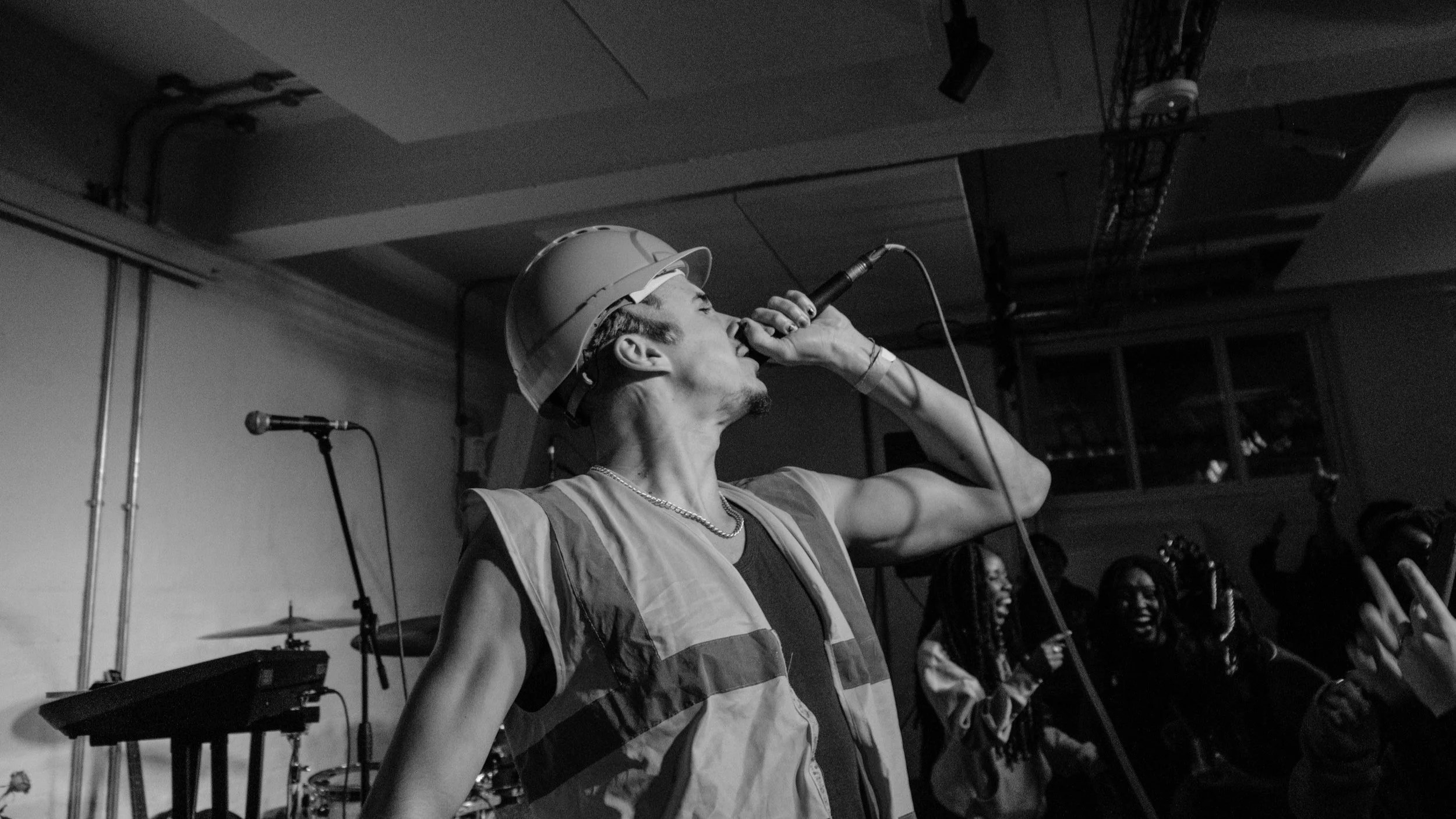 Black and white image of a person wearing a construction helmet and reflective vest, holding a microphone on stage, with crowd in the background.