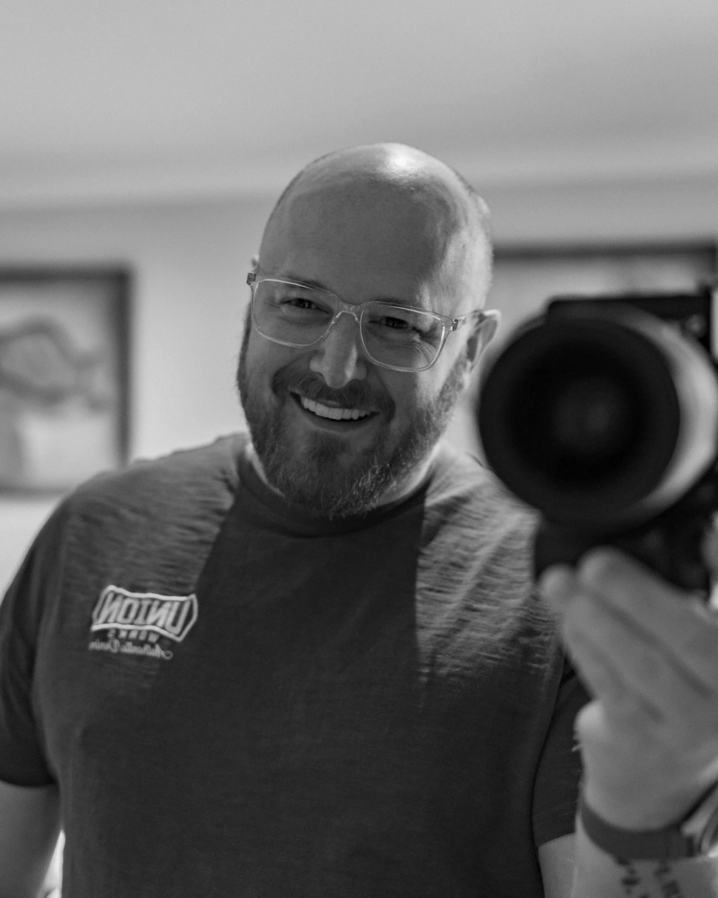 Close-up of a smiling man with glasses and a beard taking a selfie in front of a mirror, wearing a Harley Davidson shirt, in a room with framed pictures on the wall.