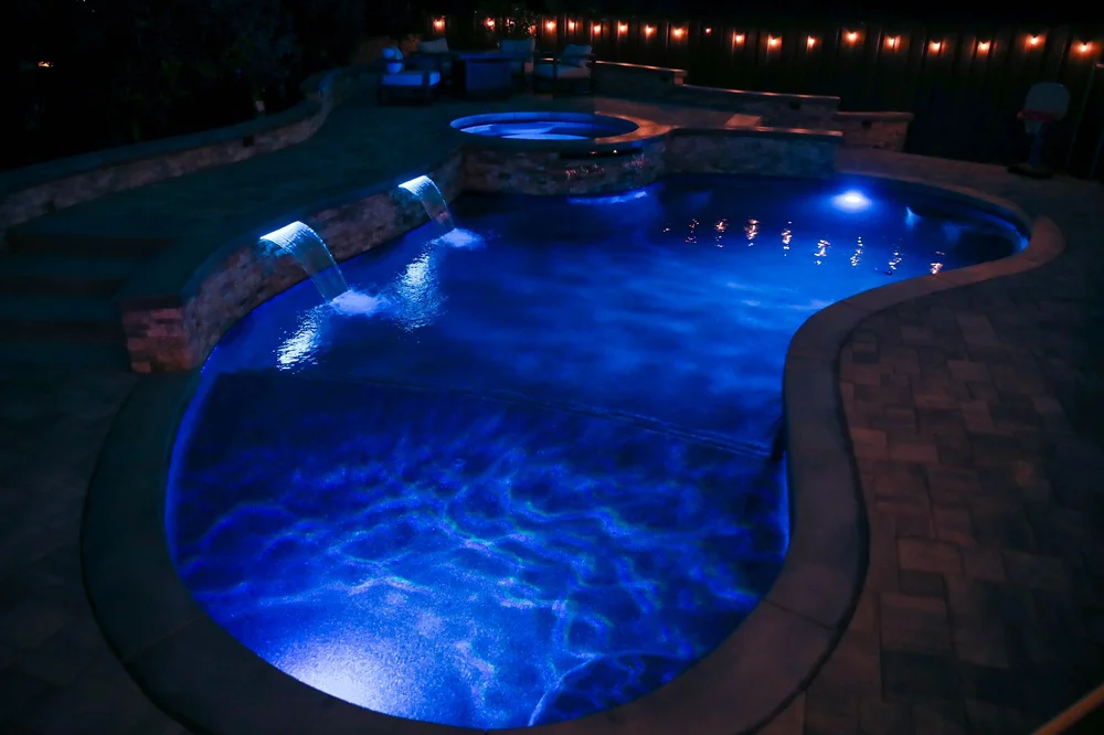 A curved pool glows blue at night beneath illuminated spillways, with a raised round spa and string lights behind it.