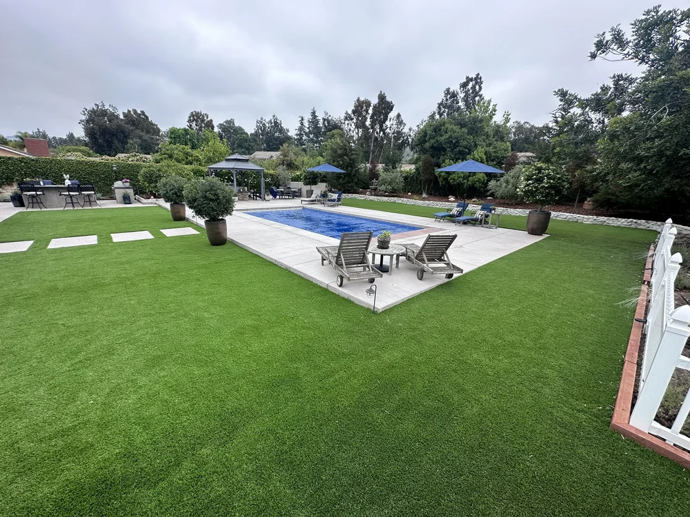 Wide garden view of a rectangular pool with a blue cover, pale deck, lounge chairs, umbrellas, and surrounding lawn.