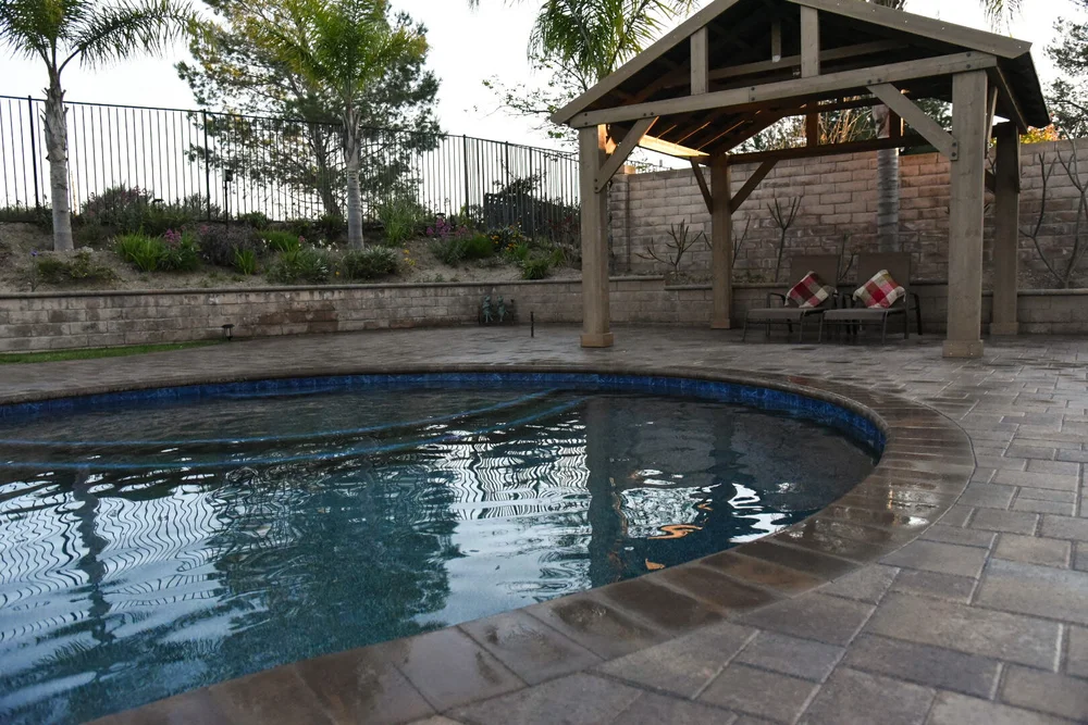 Curving pool edge and paver deck beside a timber-roofed seating area, with a planted retaining wall behind.