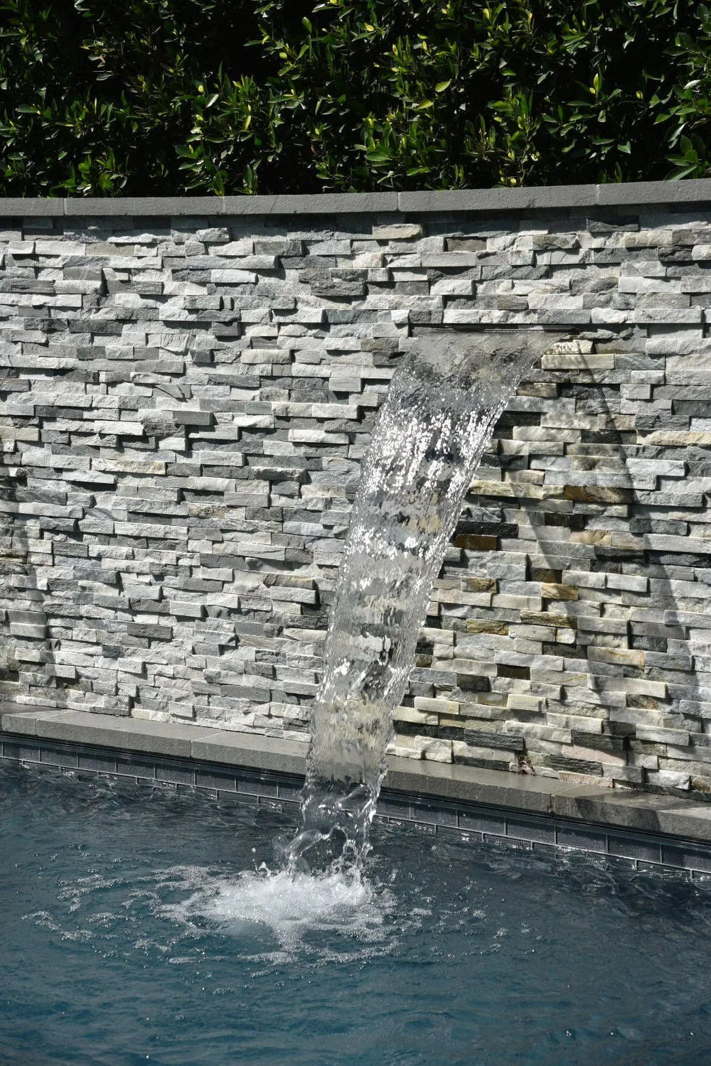 A sheet of water flowing from a narrow opening in a gray stacked-stone wall into the pool.