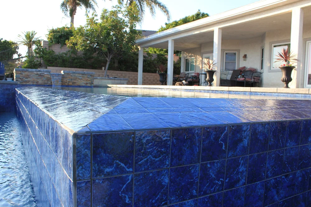 Water sheets over a broad blue-tiled pool edge, with a covered patio and stone spillway wall behind it.