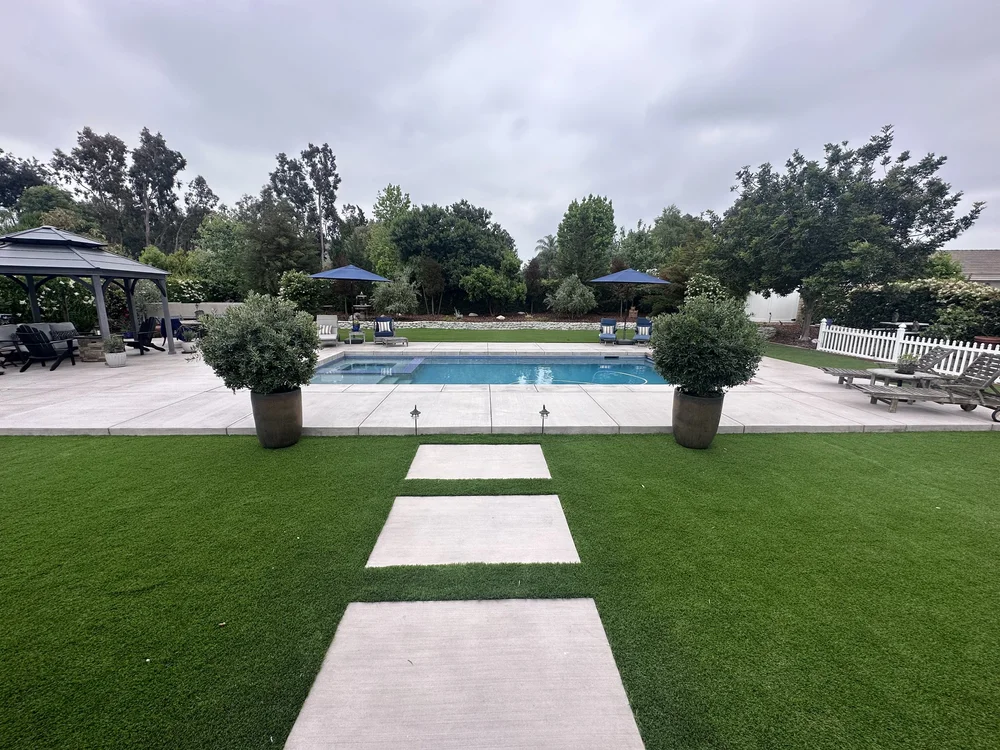 Large rectangular stepping stones cross the lawn toward a pool deck framed by matching potted trees.