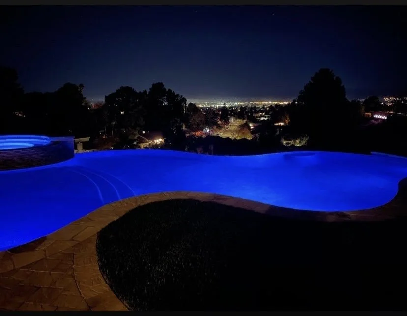 Curving pool illuminated blue at night, bordered by stone coping and a dark lawn, with distant lights beyond.