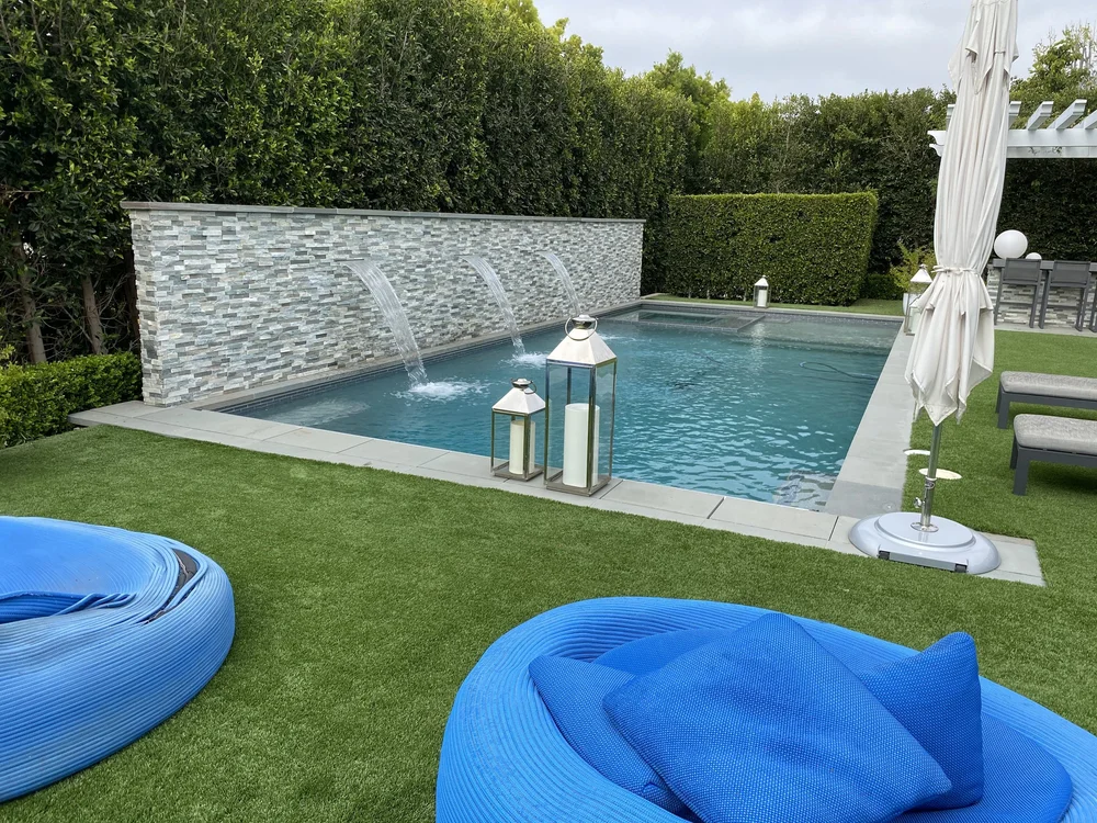 Wall-mounted streams arc into a rectangular pool beside a lawn furnished with bright blue seating and lanterns.