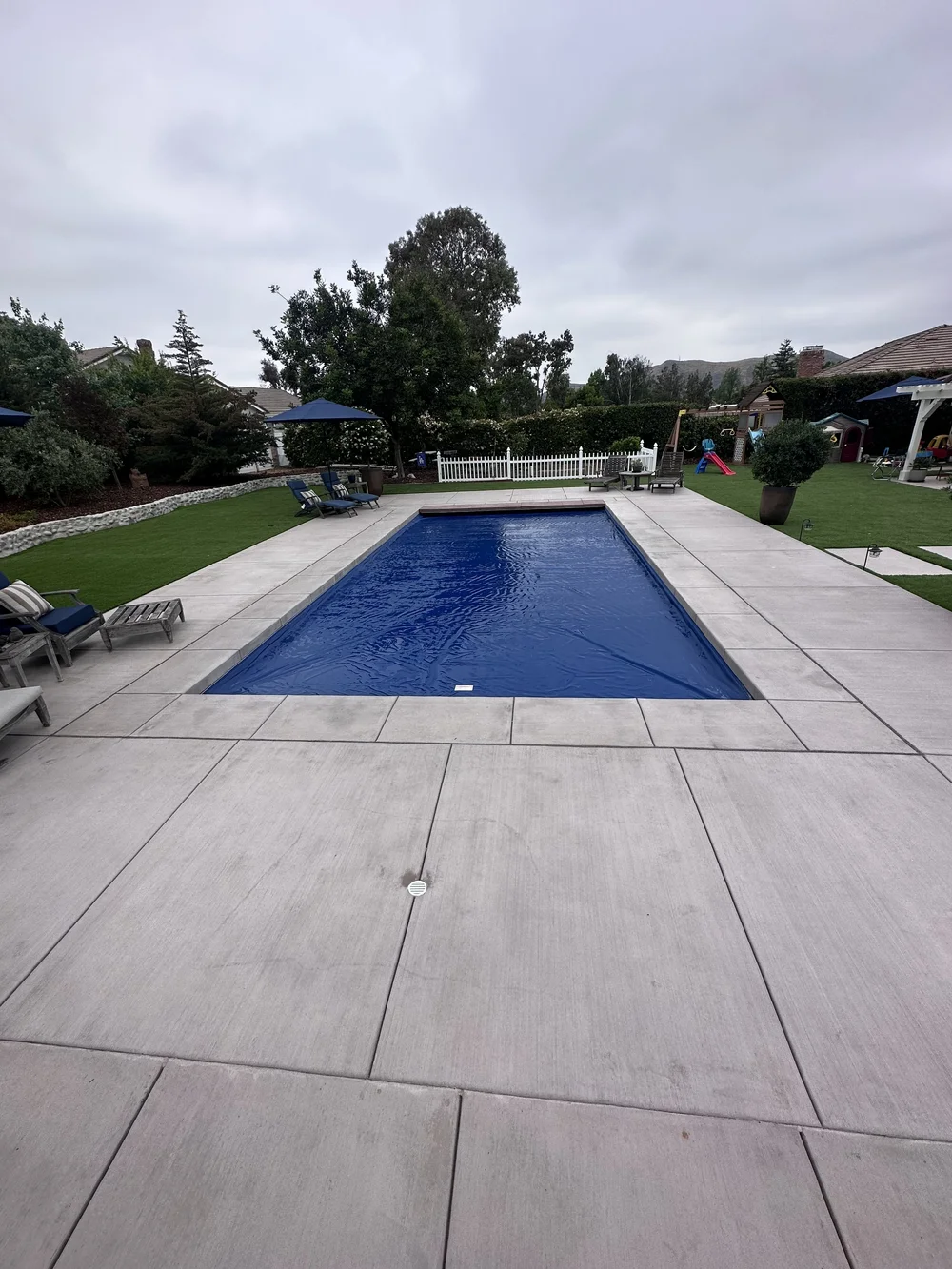 A blue cover spans a rectangular pool surrounded by a pale deck, lawn, lounge chairs, and umbrellas.
