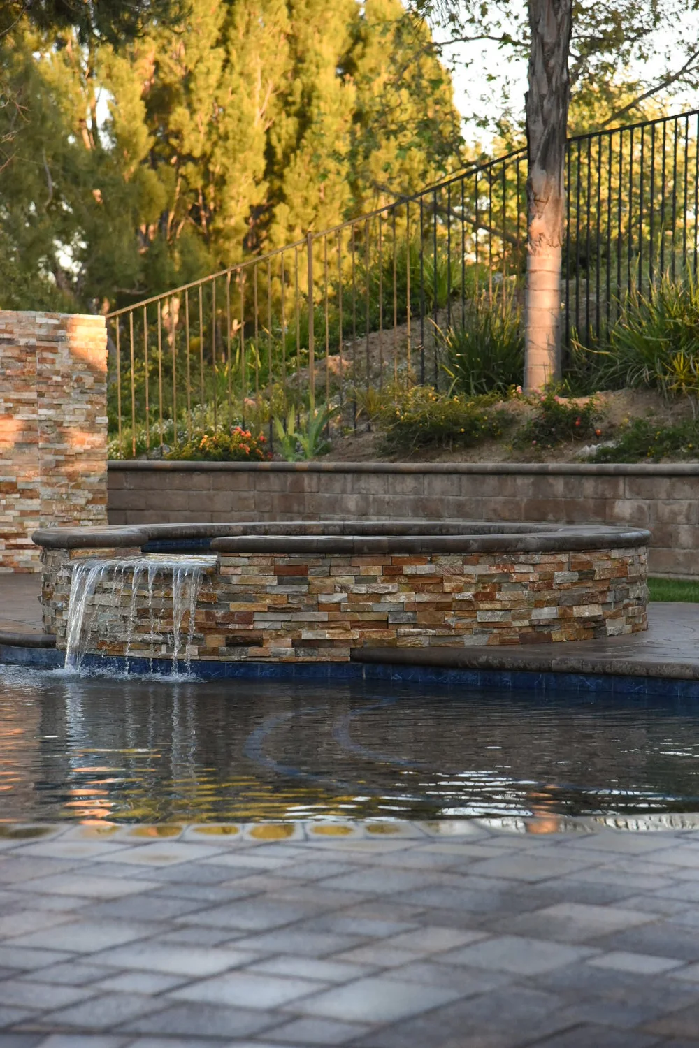 Low circular stone-faced spa with a wide spillway flowing into the pool beside a paver deck.
