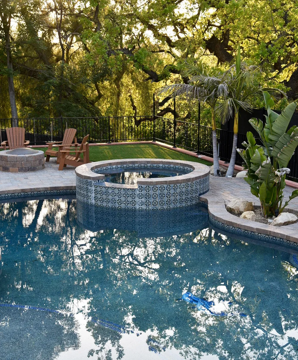 Circular tiled spa beside a freeform pool, with tree reflections, boulders and poolside seating in the background.