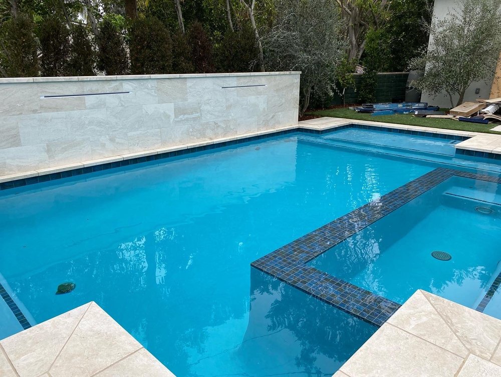 A geometric blue pool features a square inset spa edged with dark mosaic tile and a pale feature wall.