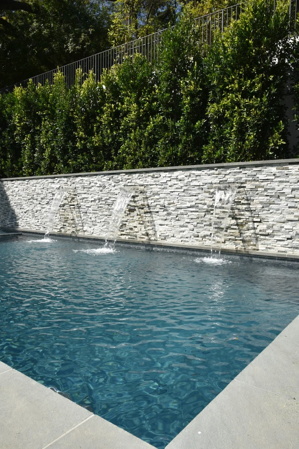 Rectangular pool with light-colored coping, a long stacked-stone water wall and a tall green hedge behind it.