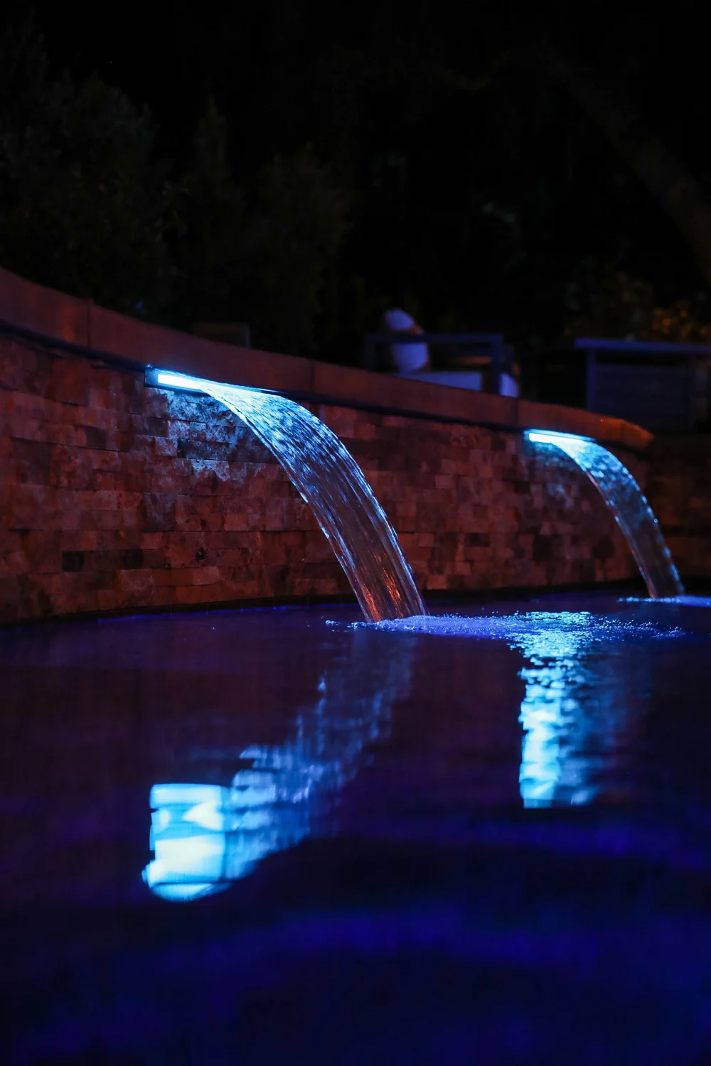Two blue-lit spillways flow from a stone wall, casting bright reflections across the pool at night.