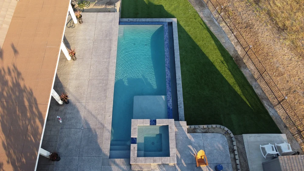 Overhead view of a narrow rectangular pool with a square spa, shallow shelf, entry steps, paved patio, and adjacent lawn.