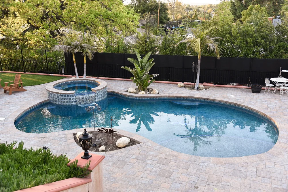 Wide view of a freeform pool and raised tiled spa, with a paver deck, boulders, palms and outdoor seating.
