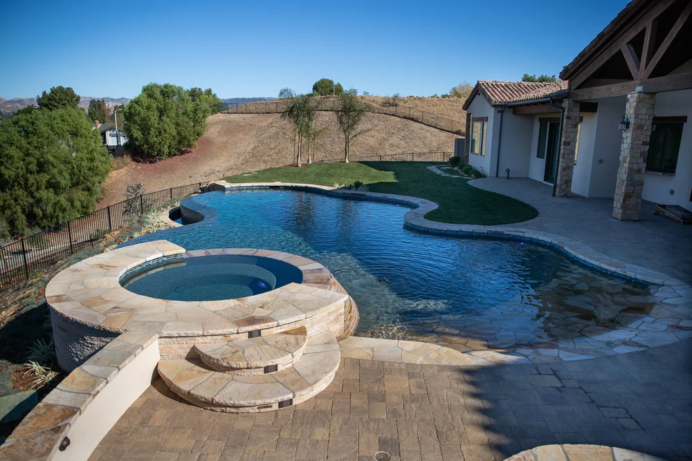 Freeform infinity-edge pool with a raised circular spa, stone surround, curved entry steps and paved patio.