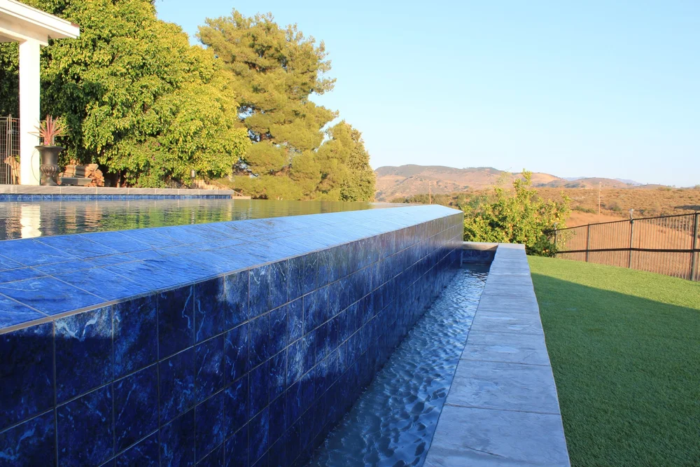 Water runs down a blue-tiled pool wall into a narrow catch basin beside a lawn and distant hills.