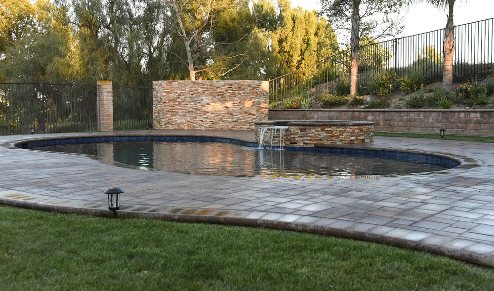 Freeform pool bordered by a paver deck and lawn, with a raised spa and stone wall across the water.