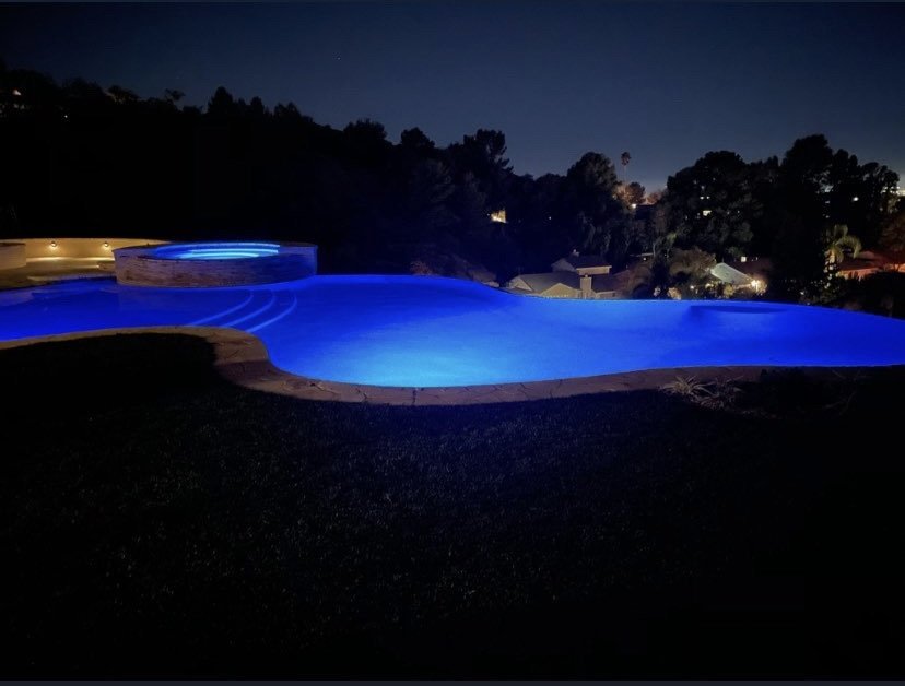 Wide nighttime view of a blue-lit freeform pool and raised circular spa surrounded by trees and lawn.