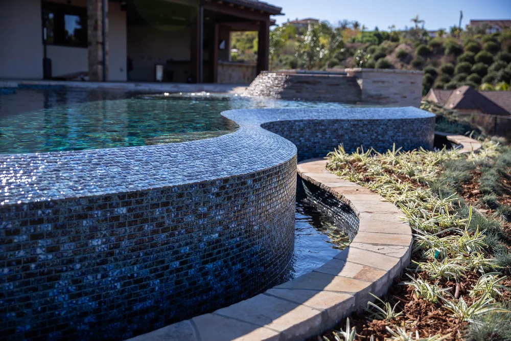 Close view of the pool's curved mosaic overflow wall and lower catch basin, with a raised spa behind it.