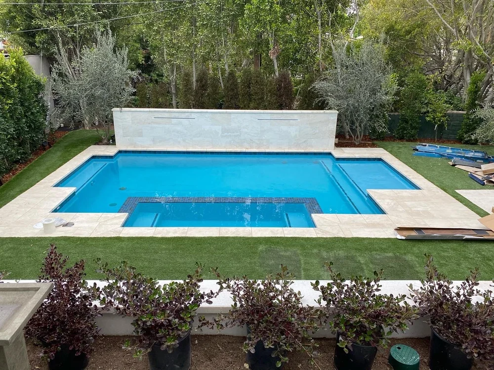 Elevated view of a geometric pool with a front inset spa, stepped shallow areas, pale coping, and a tall rear wall.