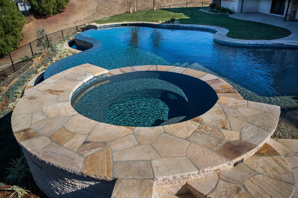 Raised round spa with a stone surround and curved entry steps beside a blue freeform pool.