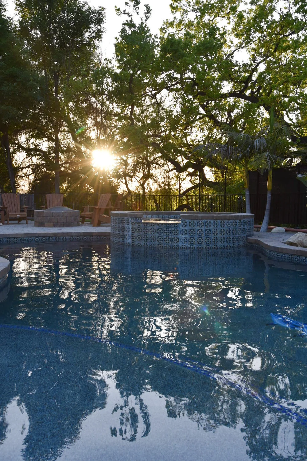 Tree silhouettes and warm evening sunlight reflected in a freeform pool with a raised circular spa.